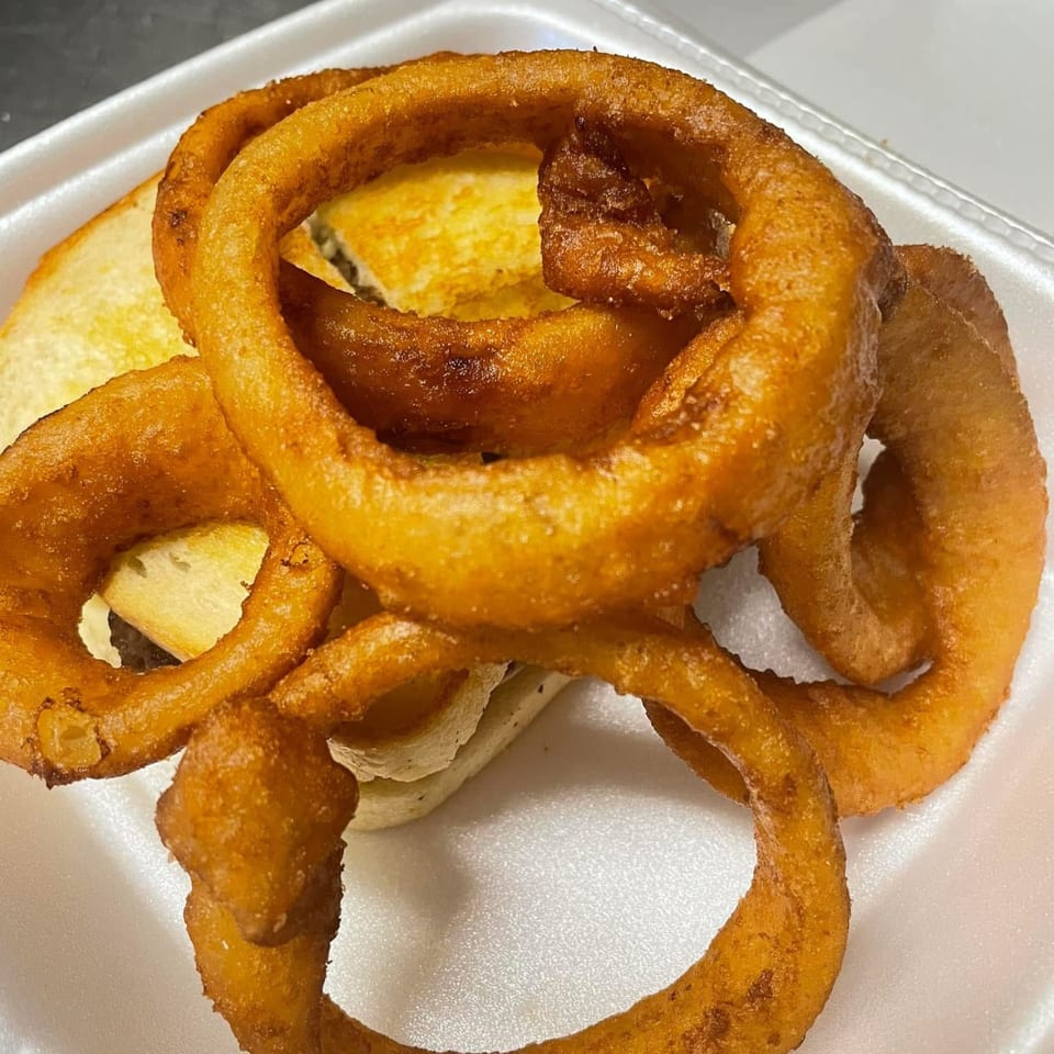 Golden-brown, crispy onion rings piled on top of a burger in a white takeout container.